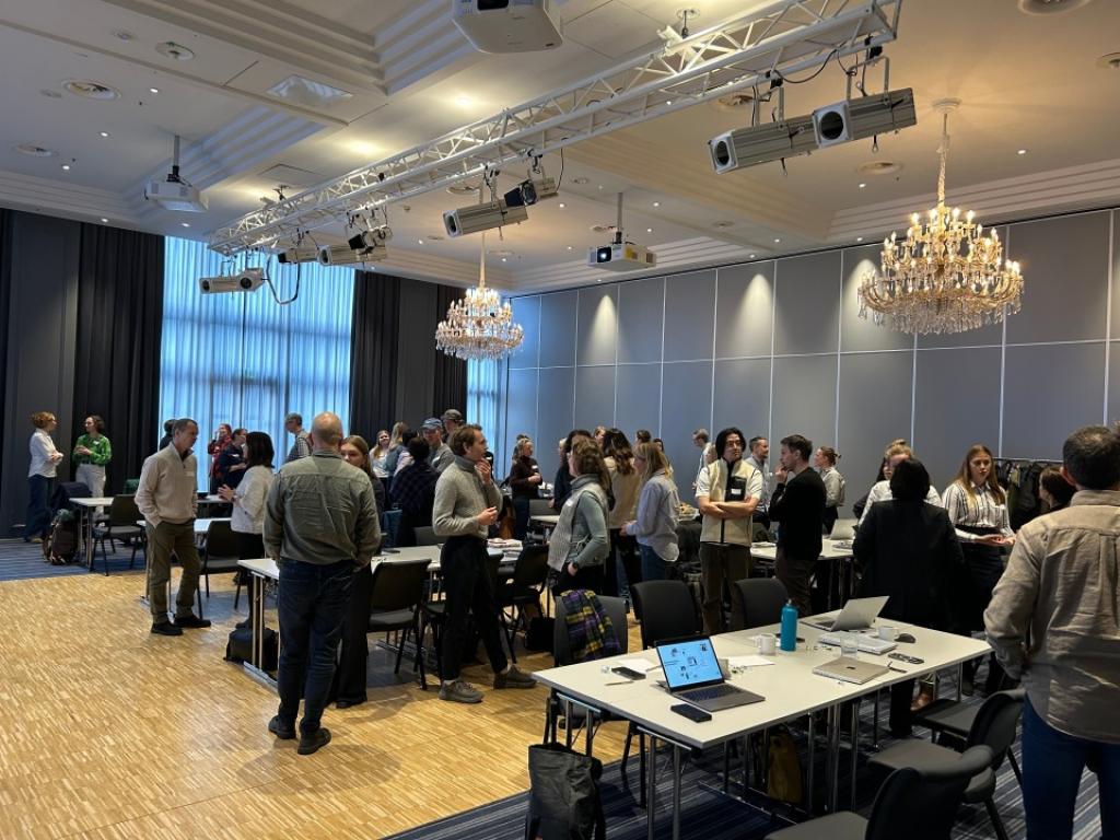 Mange deltakere står i grupper og snakker sammen i en konferansesal.