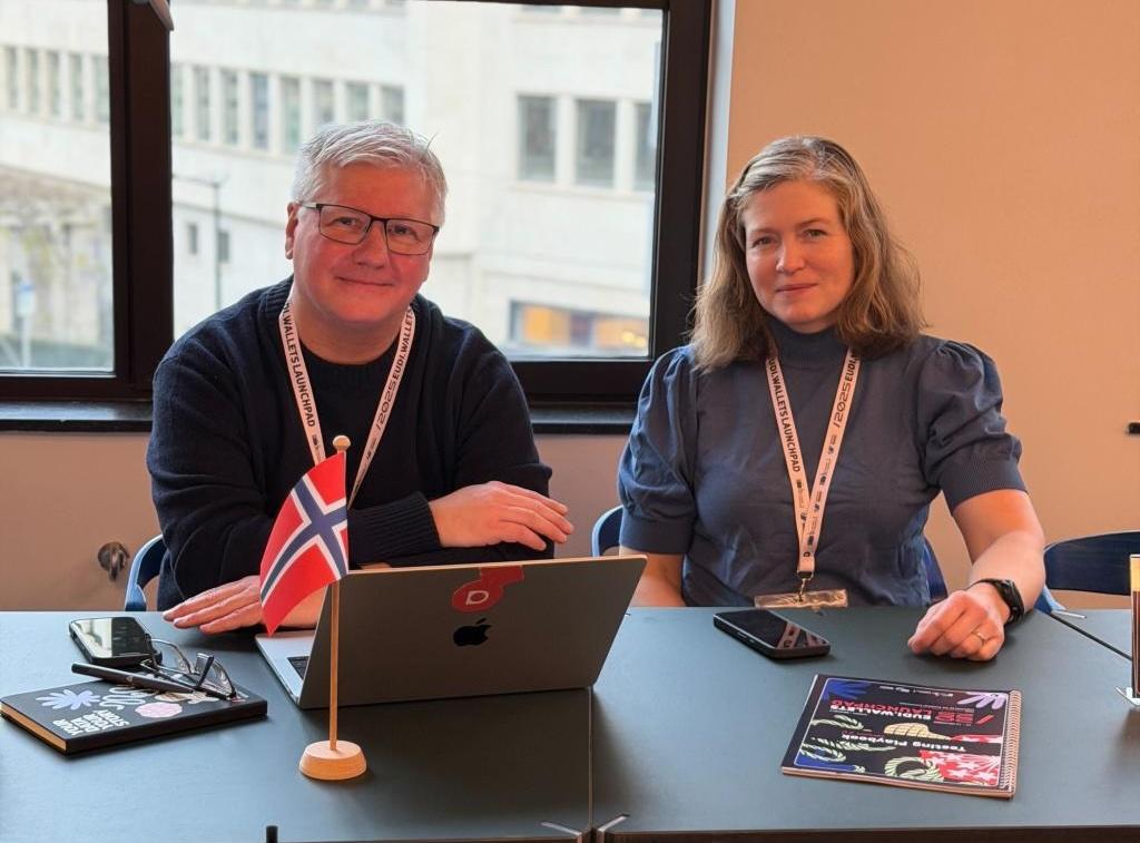 Thomas og Randi smiler til kamera, der de sitter bak bordet med pc-en foran seg og norgesflagg